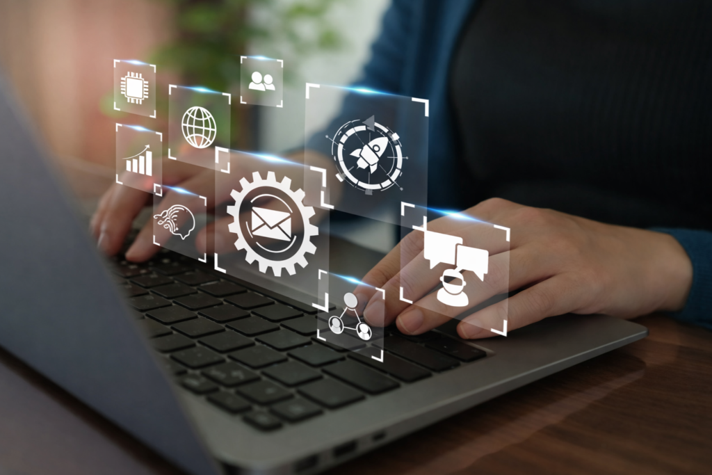 8 AI Skills 2026 That Actually Matter (Not Just Hype) 4 Close-up of hands typing on a laptop with floating holographic icons representing automation, global connectivity, analytics, messaging systems, and AI-driven business workflows in a modern office environment.