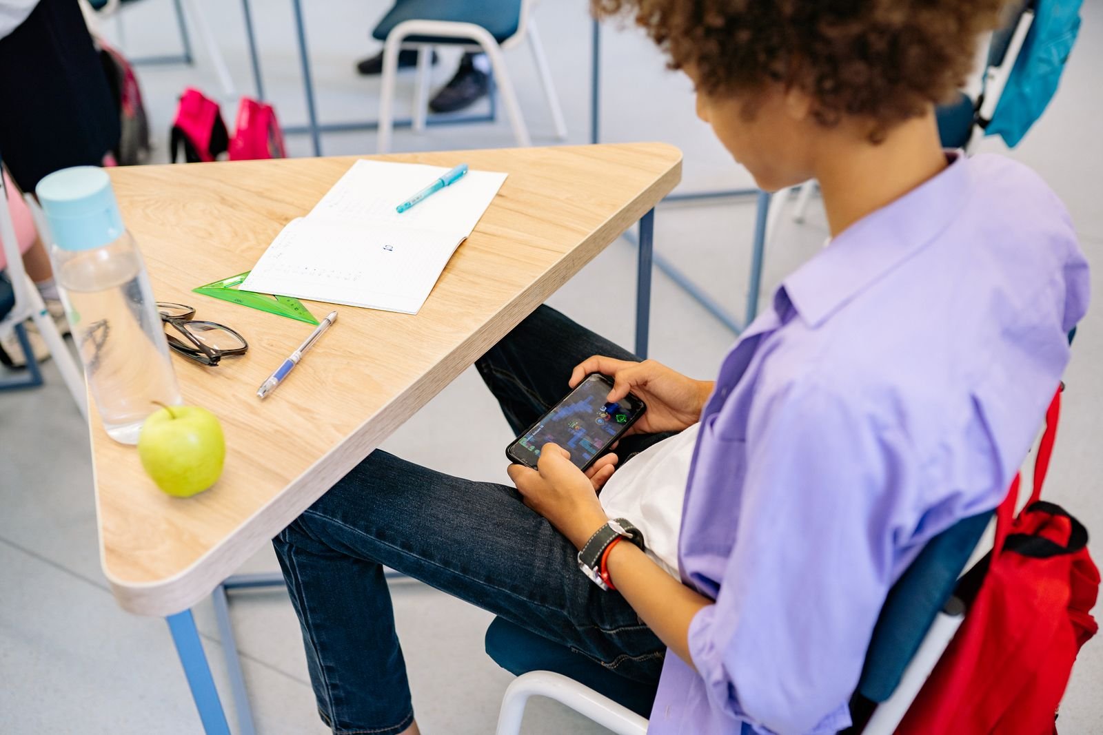 Gemini in Classroom Just Opened to K-12. There's No Parent Off Switch. 5 A student holding a smartphone below a school desk beside a backpack