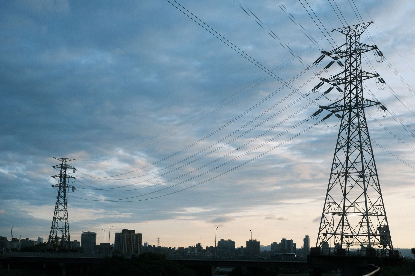 Transmission towers and power lines silhouetted over a city skyline at dusk, the infrastructure behind AI electricity prices